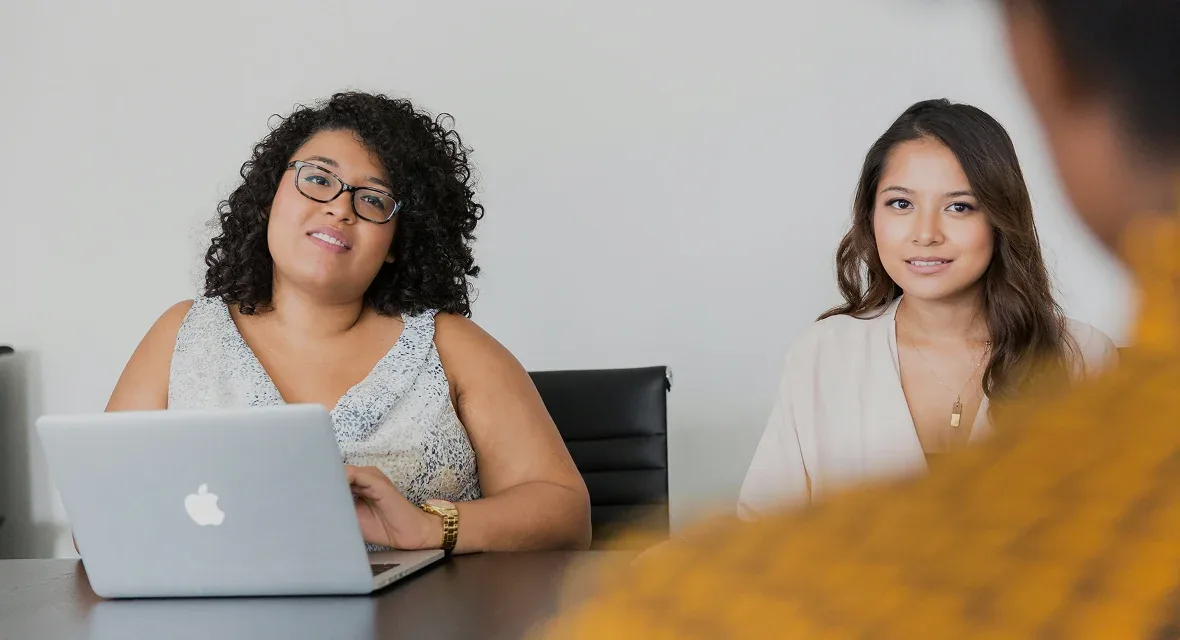 Dos mujeres sonríen sentadas a una mesa con un portátil